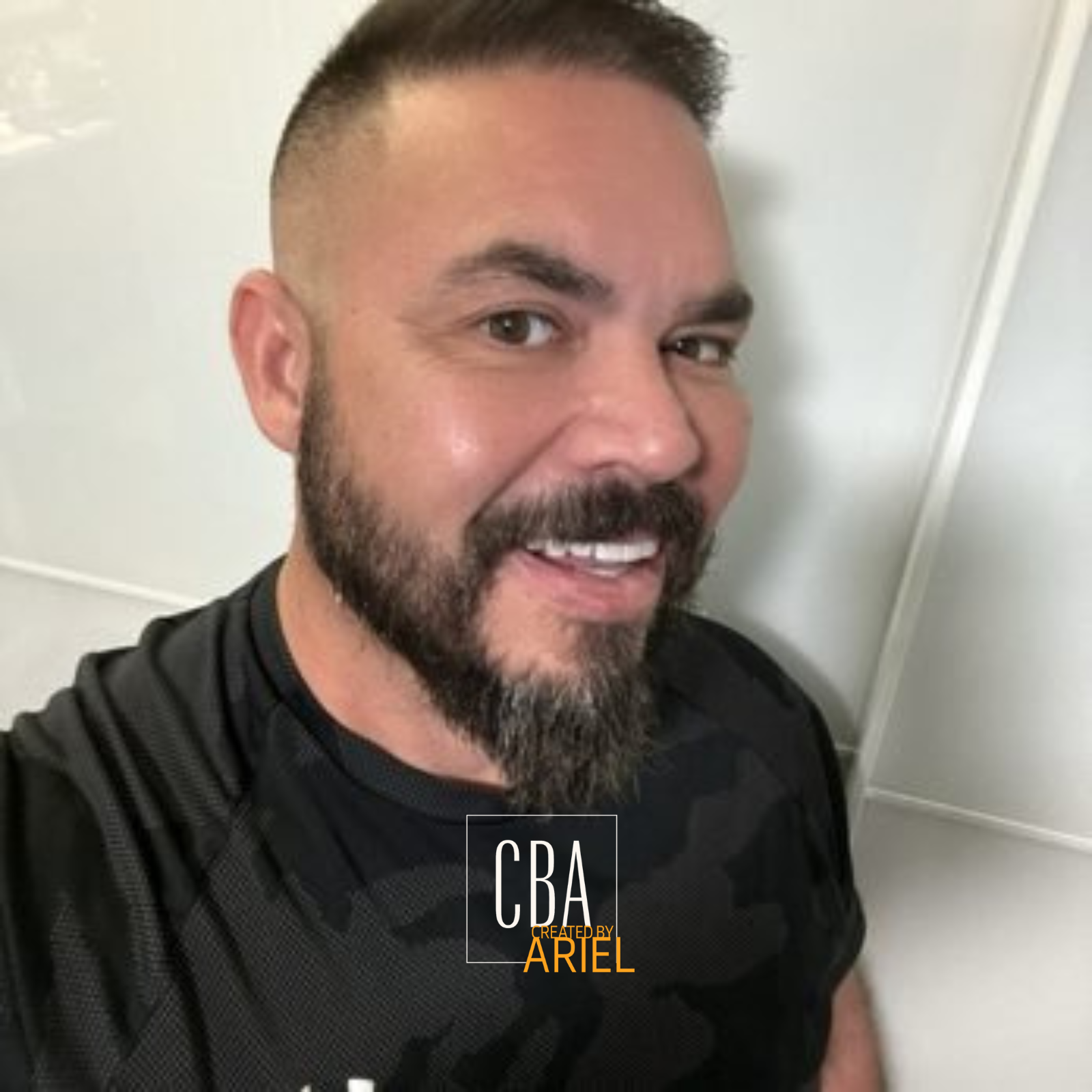 A smiling man with short dark hair, a beard, and light skin taking a selfie in front of a white wall.