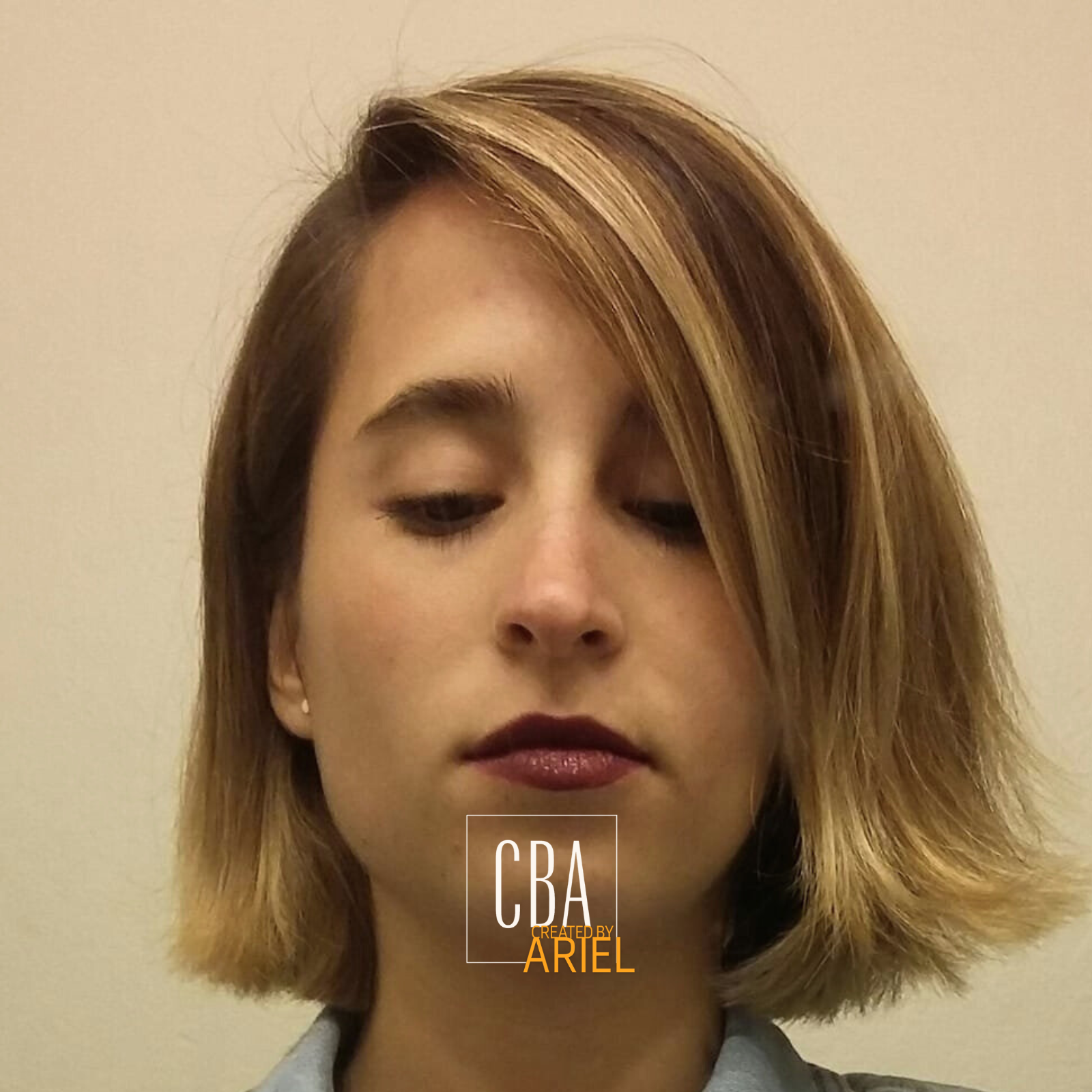 A young woman with chin-length blonde hair and side-swept bangs, looking down, with a neutral expression. She is wearing a light-colored top and small earrings, standing against a plain, light-colored background.