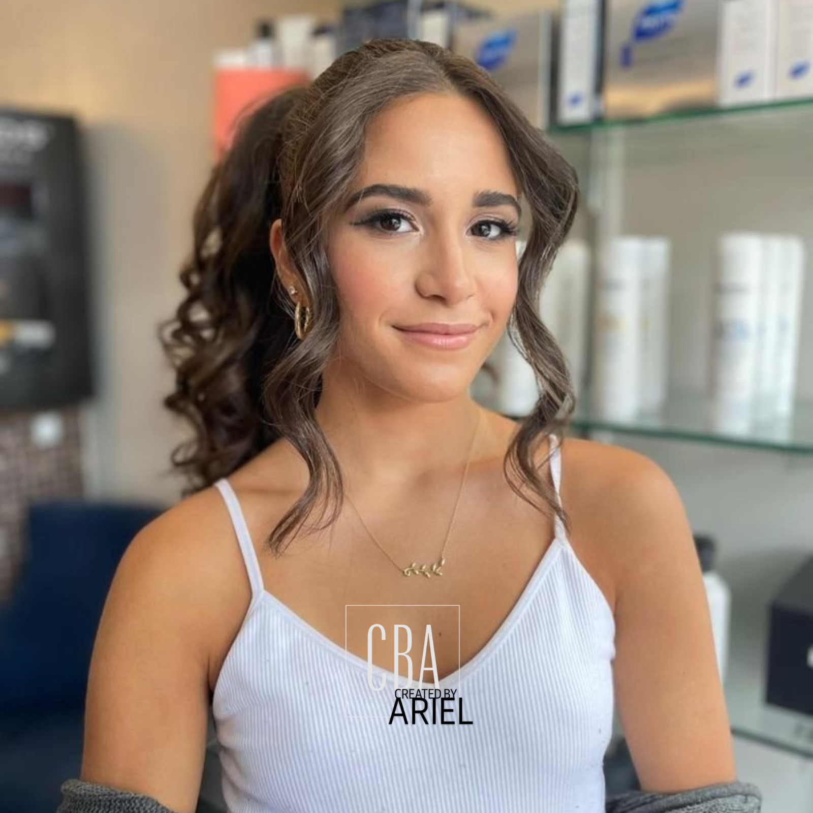 A young woman with wavy brown hair, wearing gold hoop earrings and a delicate gold necklace, in a casual white tank top, sitting in a modern salon or beauty store with shelves and hair care products visible in the background.