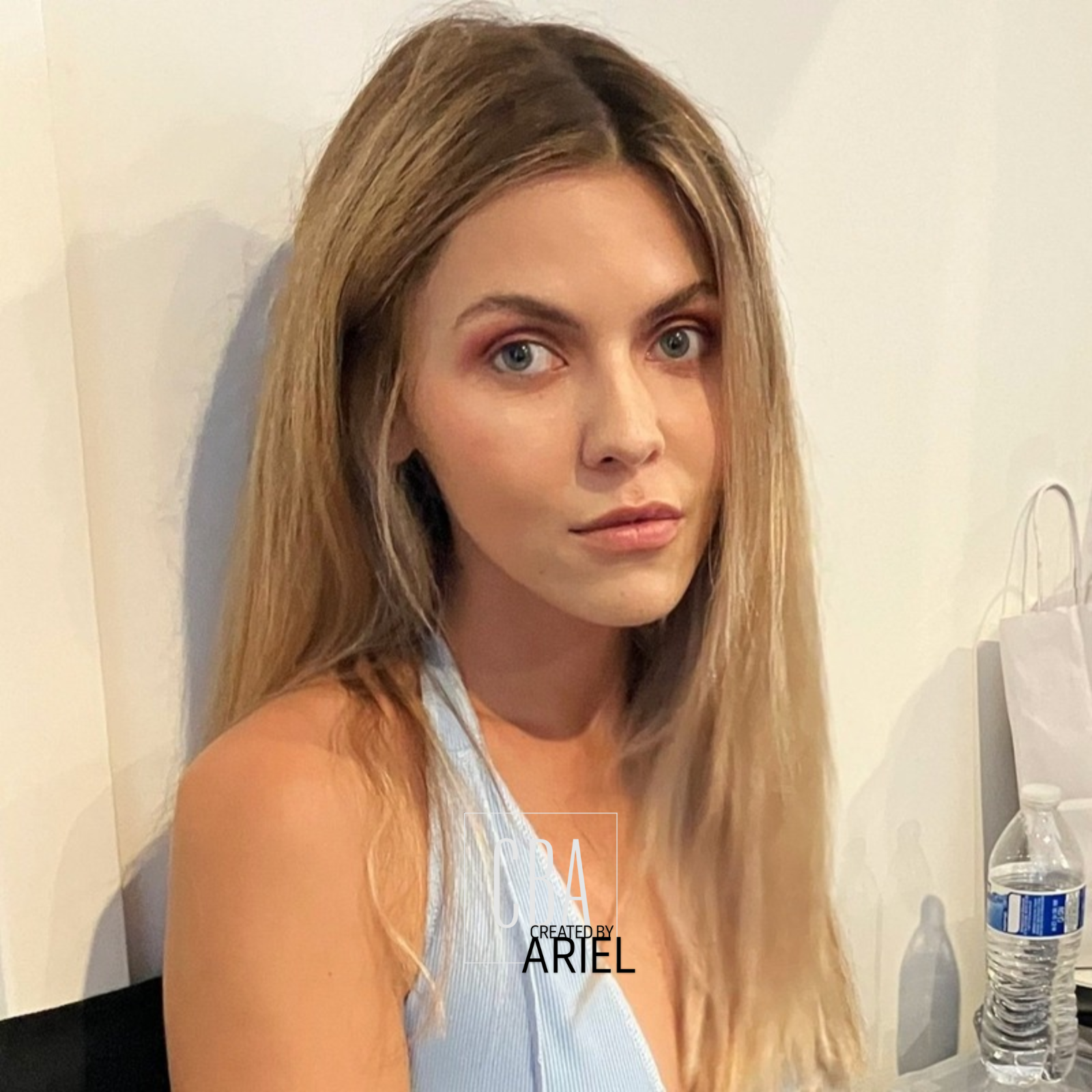 A young woman with long, light brown hair and blue eyes sitting indoors, looking directly at the camera, with a white wall in the background and a water bottle on a table beside her.