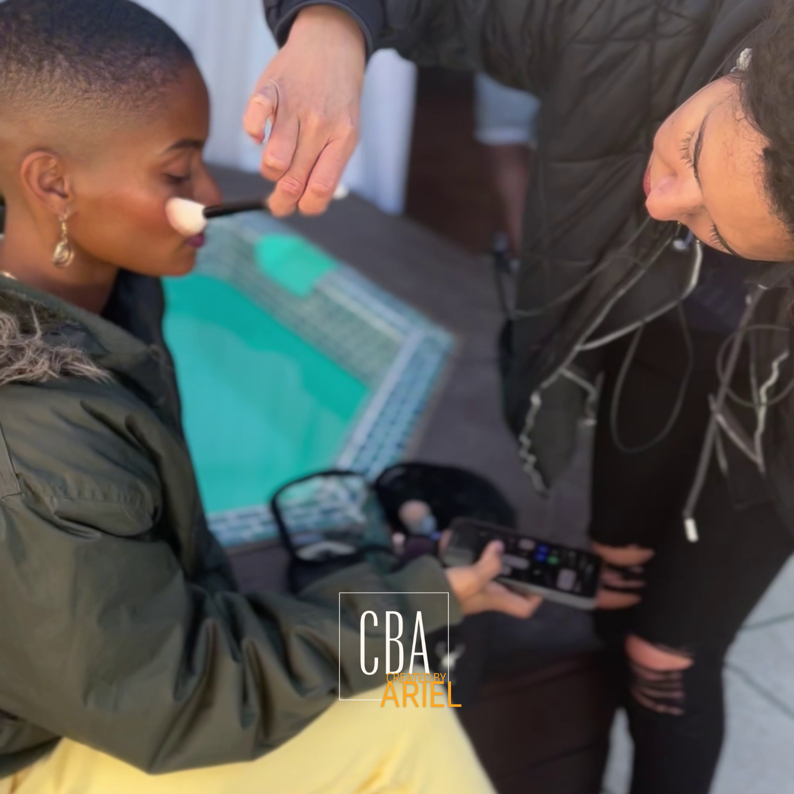 A woman applying makeup to a seated woman with a brush. The seated woman has earrings and is wearing a jacket. Another person standing is looking at a phone and holding a makeup palette. The scene is indoors with a blurred background.