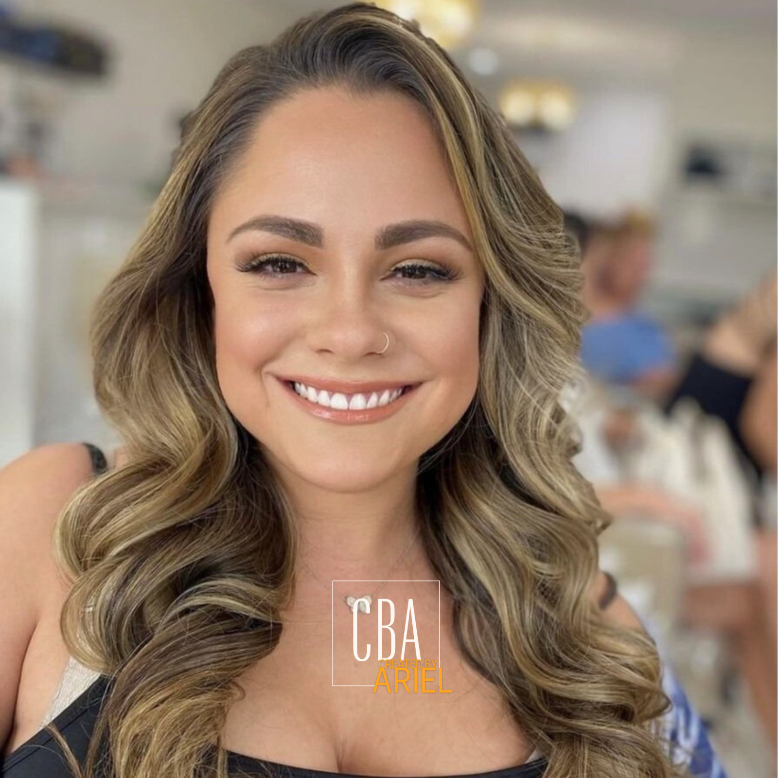 A smiling woman with long, wavy blonde hair, wearing makeup and a nose ring, in a busy indoor setting.