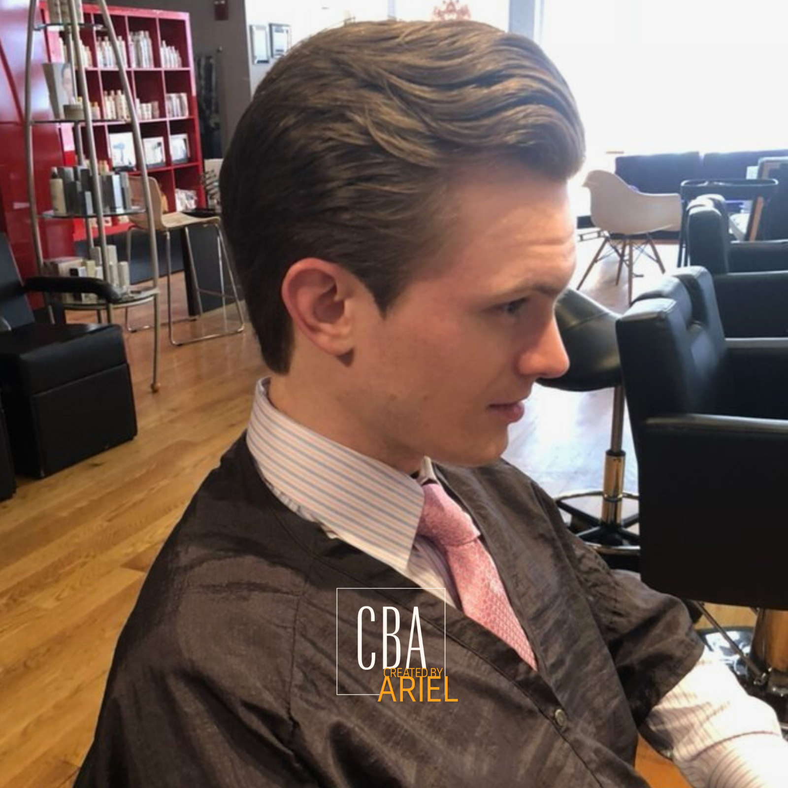 Side profile of a young man with styled brown hair, wearing a striped shirt, pink tie, and black jacket in a lounge or office space with chairs, bookshelves, and wooden floors.