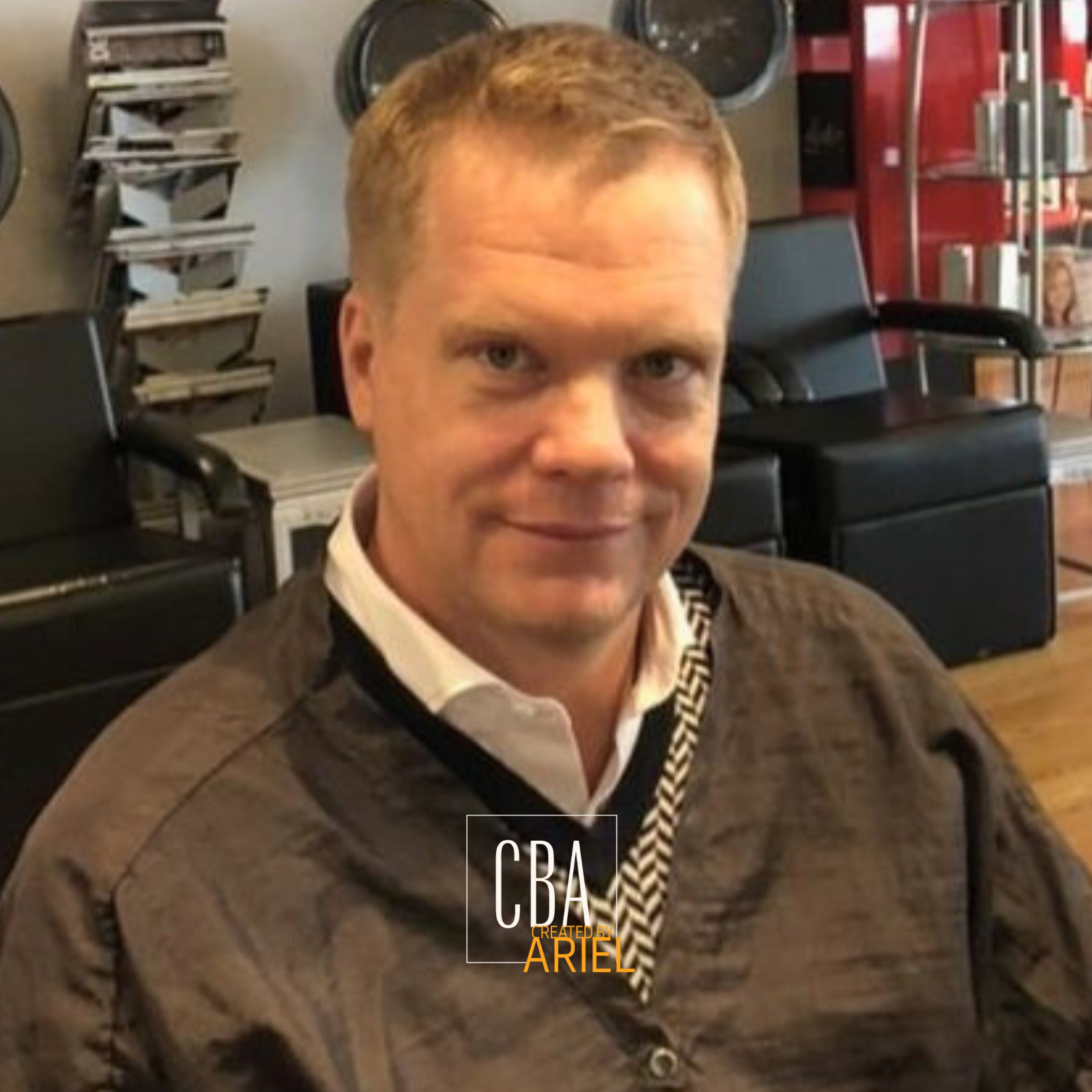A smiling man with short, light brown hair, wearing a white collared shirt and a brown jacket, sitting in a salon with hair styling chairs and tools visible in the background.