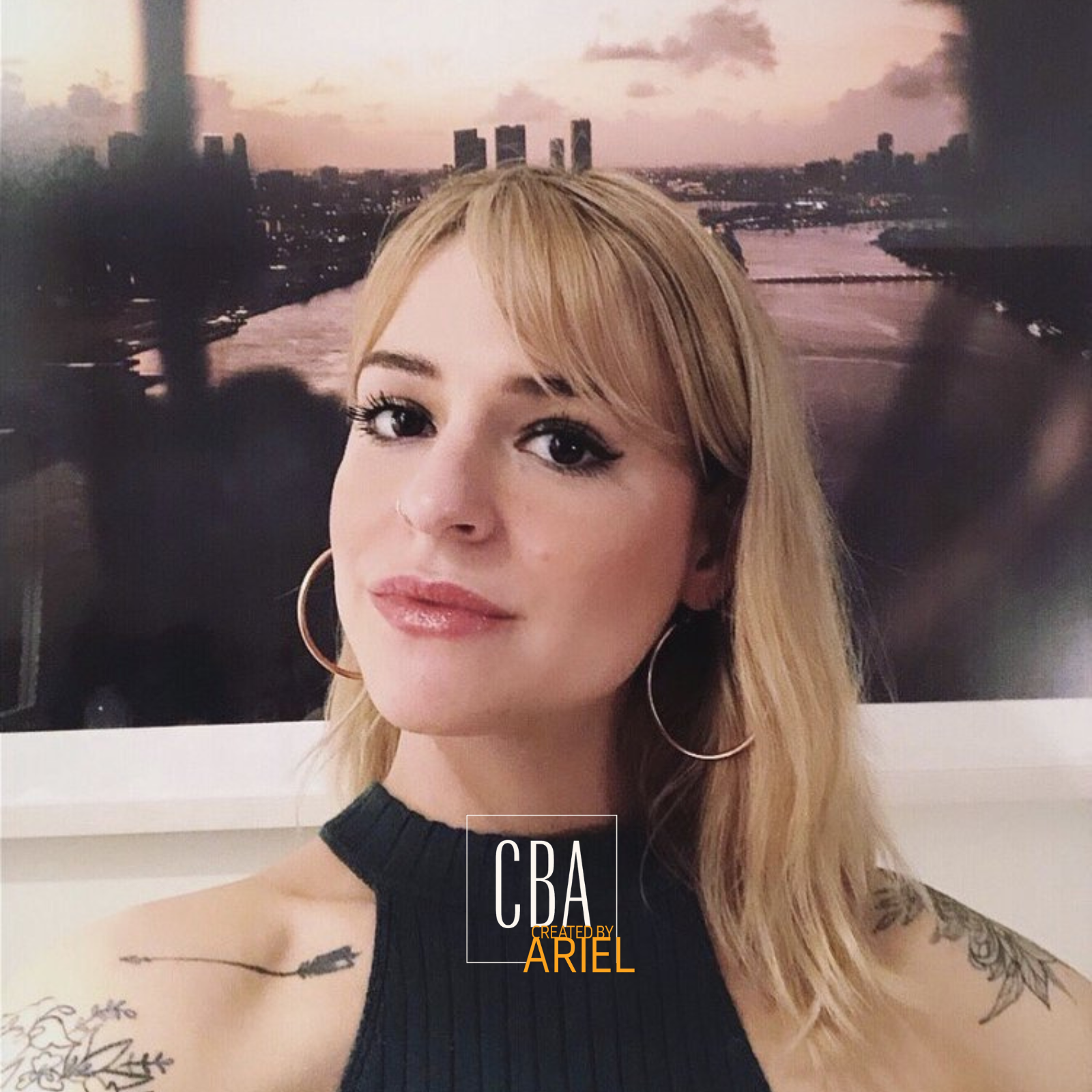 A woman with blonde hair, wearing large hoop earrings, a black sleeveless top, and tattoos on her shoulders, standing in front of a cityscape with a river and tall buildings at sunset.