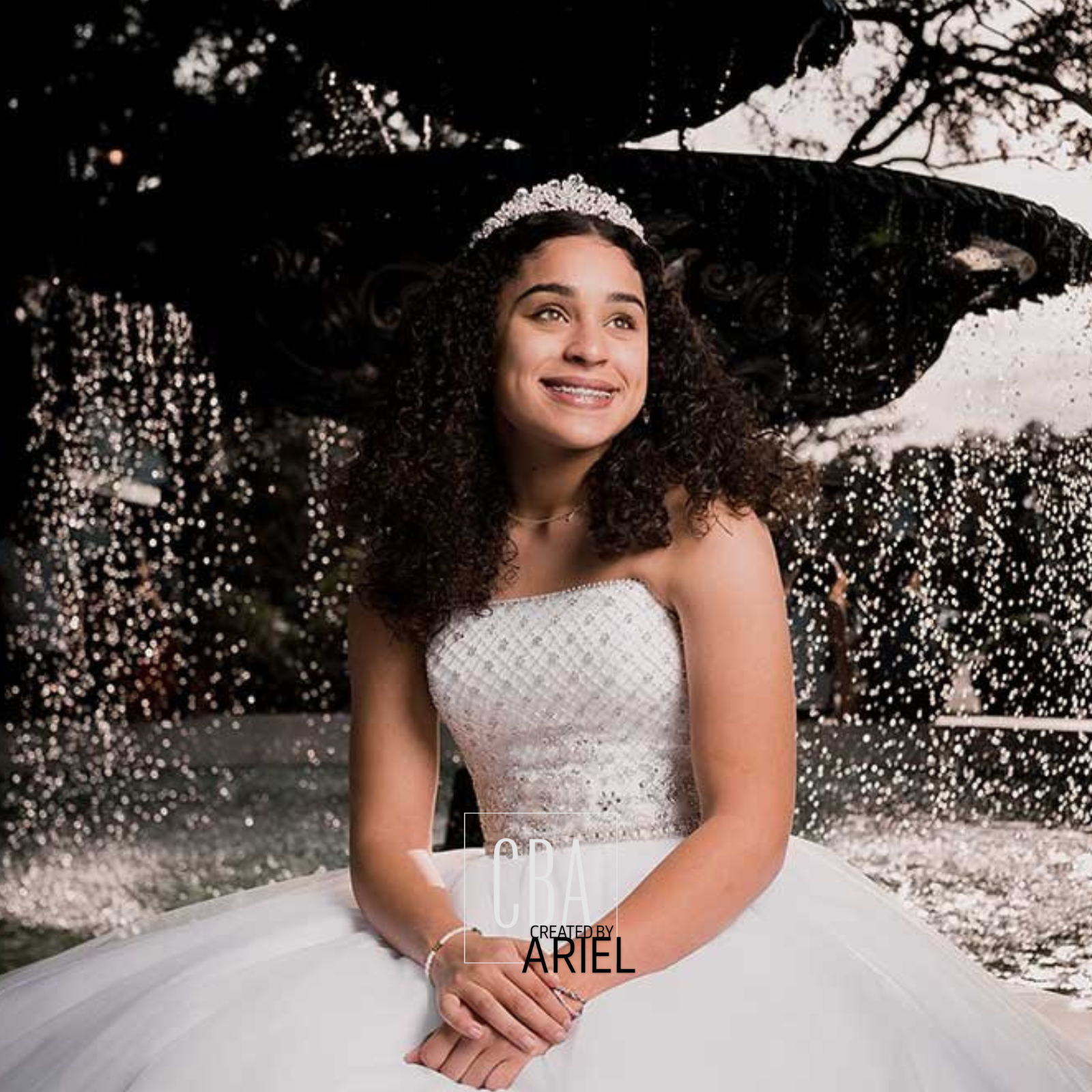 Young woman in a white strapless dress with a tiara, smiling, sitting outside under dark trees with sparkling lights in the background.