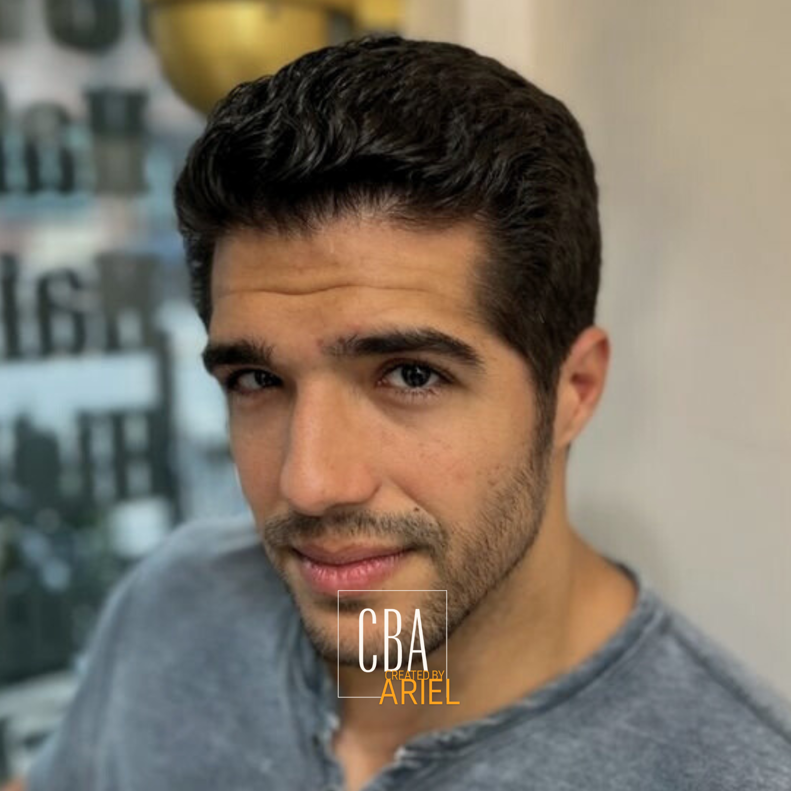 Close-up of a young man with dark, wavy hair and a short beard, wearing a gray shirt, looking at the camera with a slight smile, with blurred background.