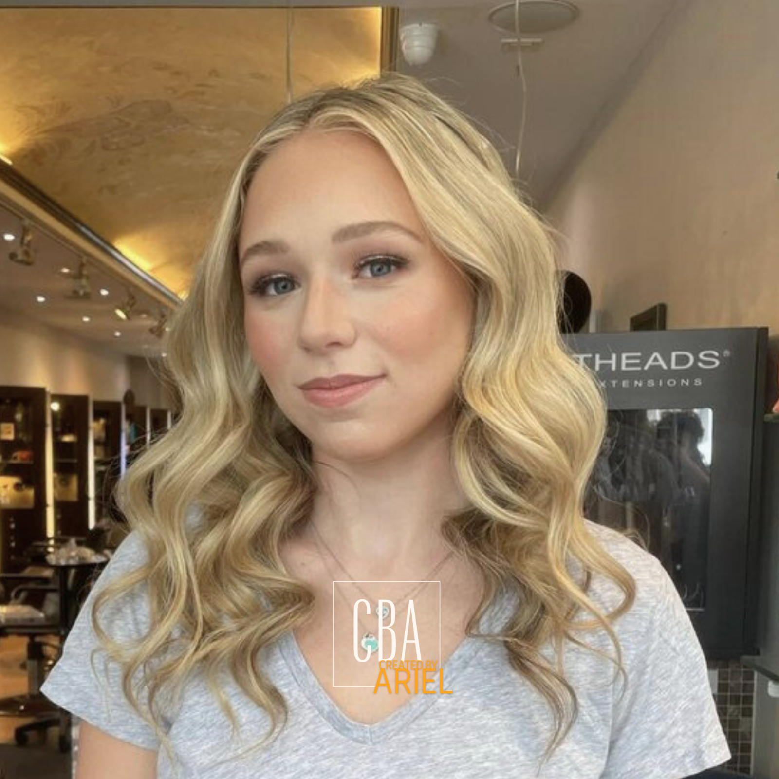 A young woman with long wavy blonde hair and light makeup, wearing a light gray T-shirt, posing inside a hair salon with wooden shelves and salon equipment in the background.
