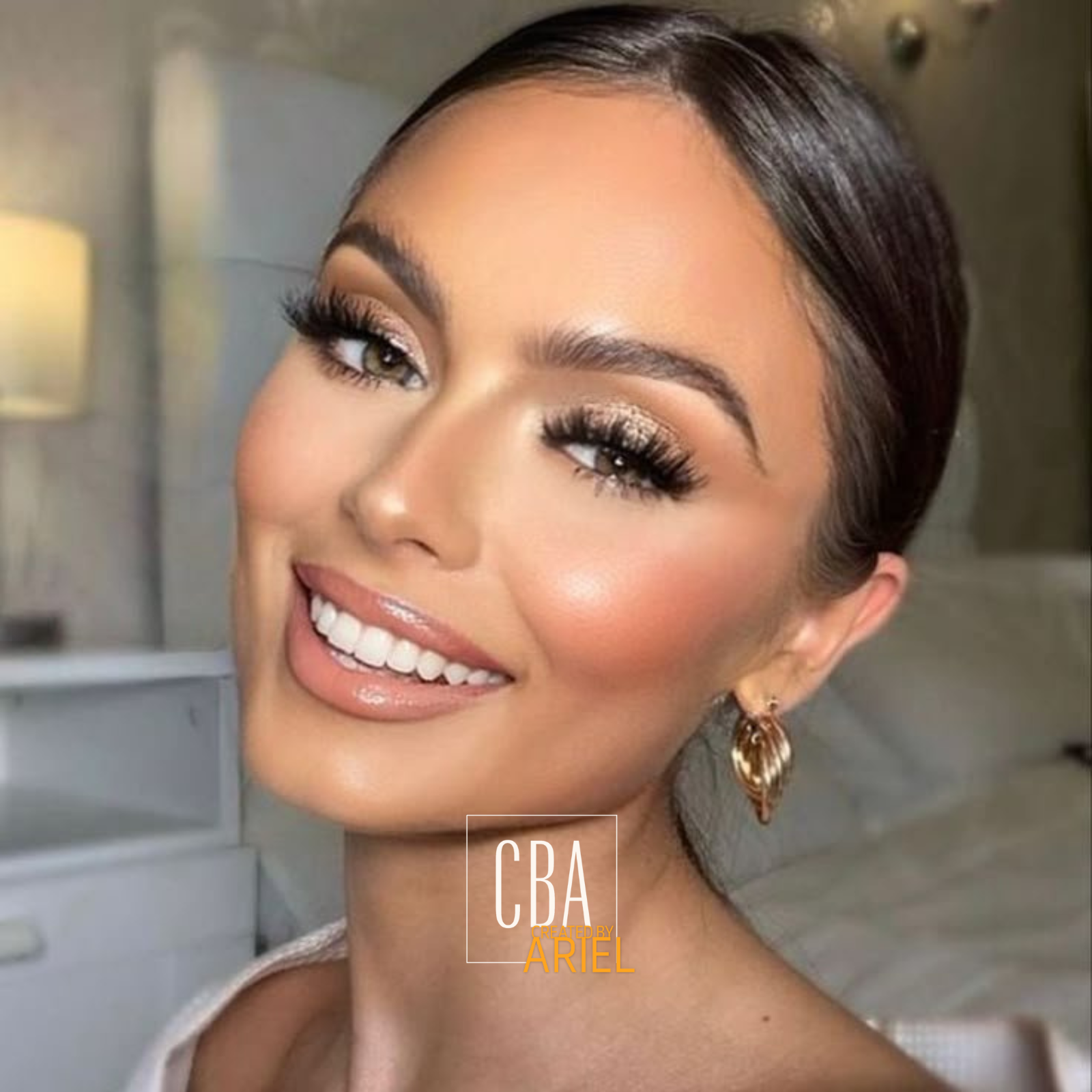 Close-up of a smiling woman with makeup, earrings, and short dark hair, in a soft-lit indoor setting.