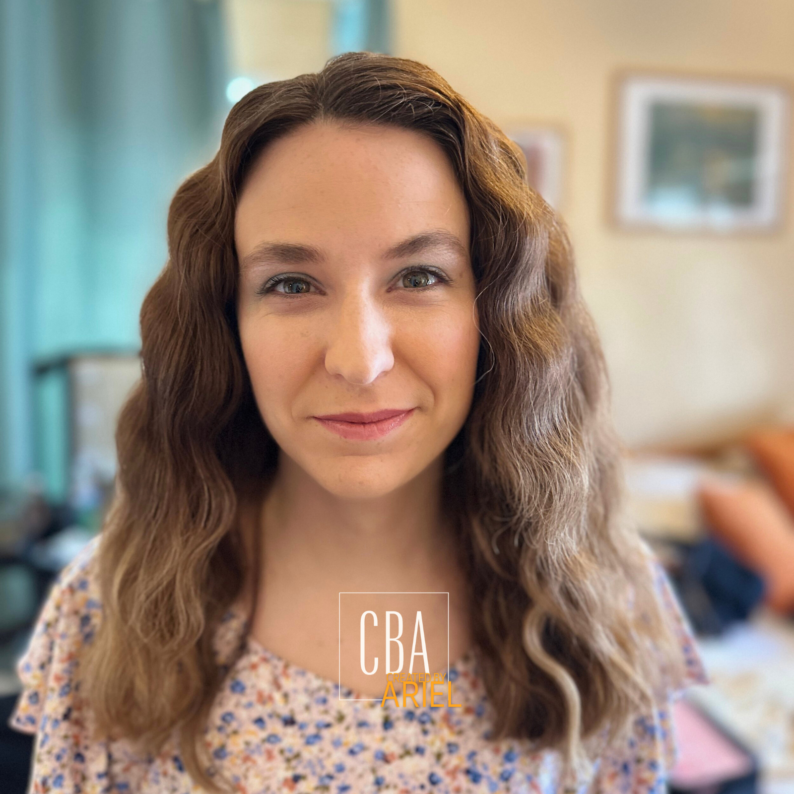 Close-up of a woman with wavy brown hair, smiling softly, in an indoor setting with blurred background.