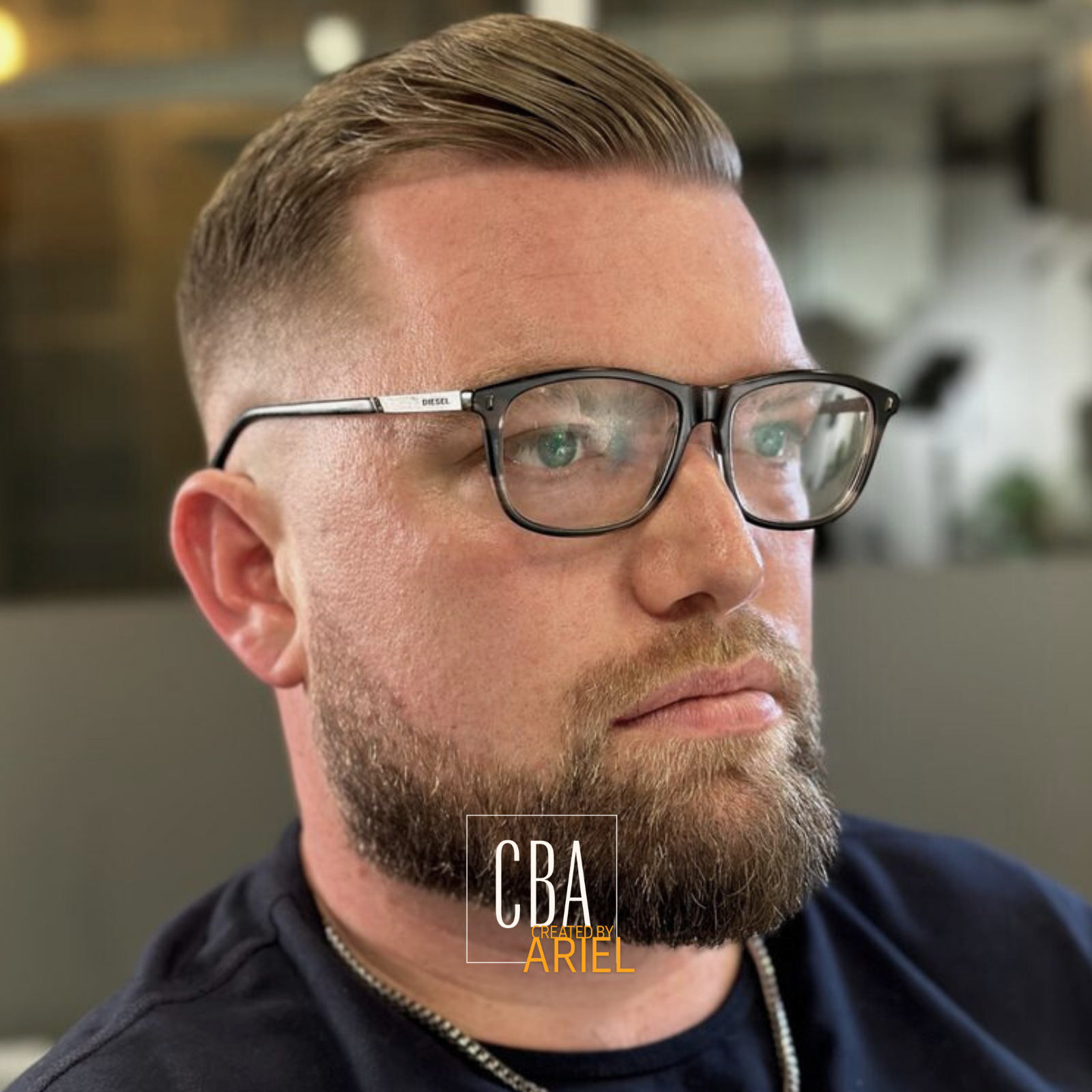 Close-up of a man with short, slicked-back hair, beard, wearing glasses and a black shirt, looking to the right, with a blurred background.