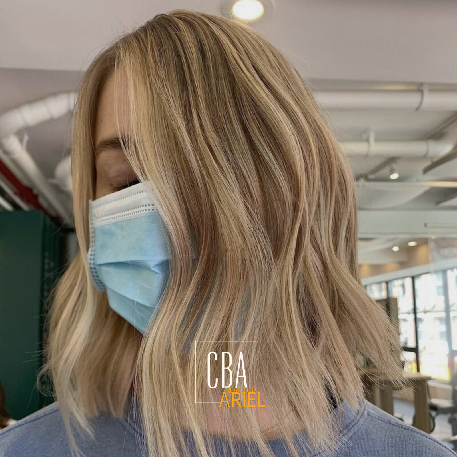 A woman with wavy blonde hair wearing a light blue surgical mask and a gray shirt, indoors in a modern setting with large windows and various ceiling fixtures.