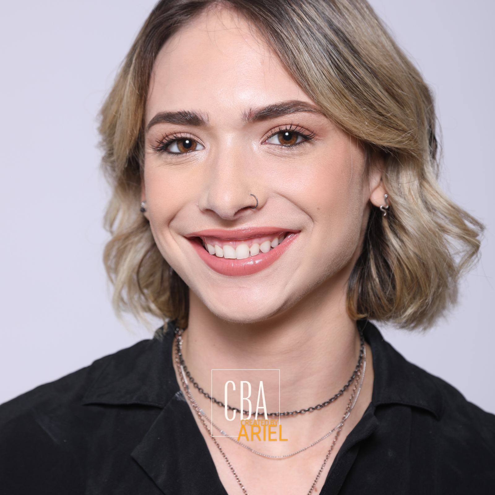A young woman with short, wavy blonde hair smiling at the camera, wearing earrings, a nose ring, layered necklaces, and a black shirt, against a light grey background.