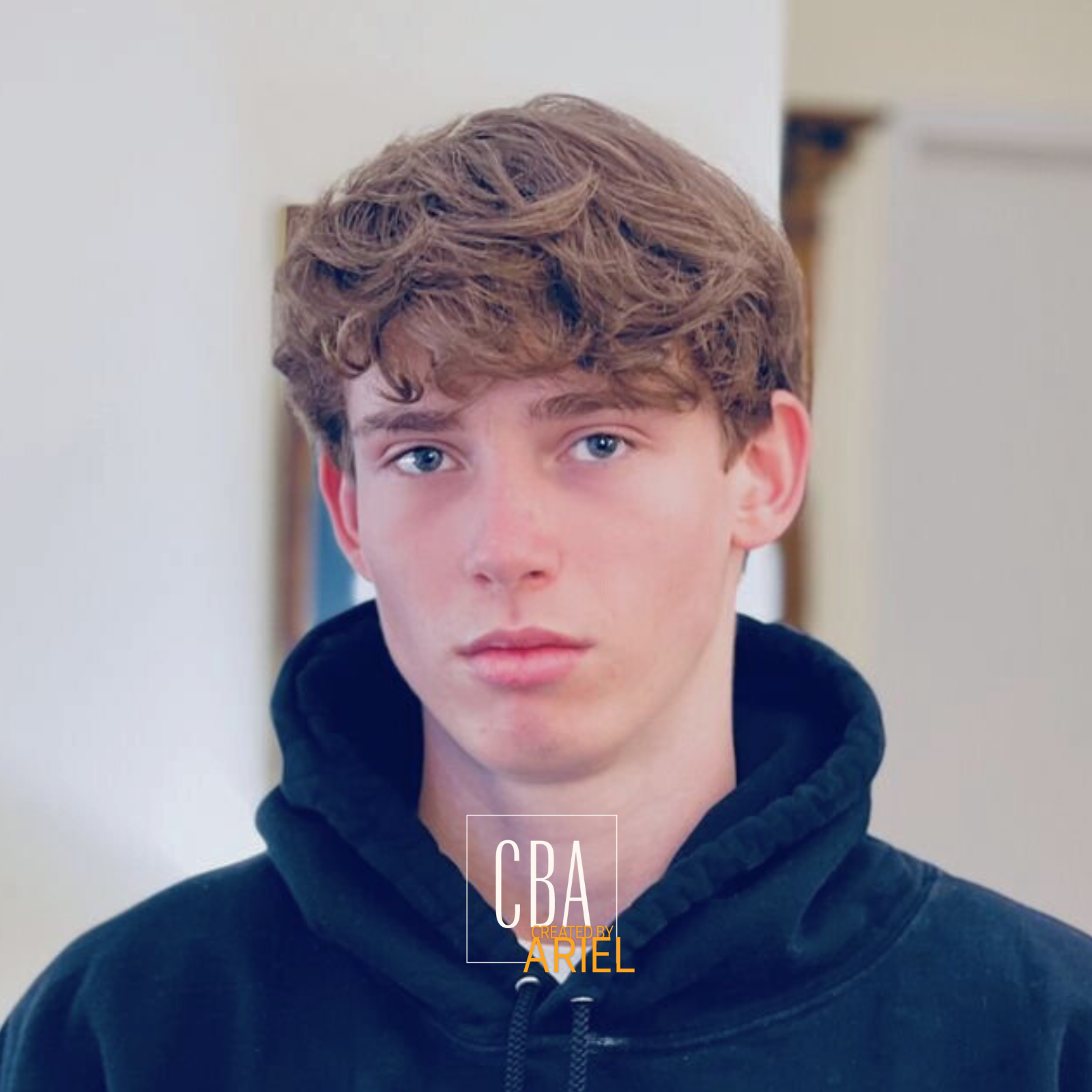 Portrait of a young man with light brown, curly hair, blue eyes, and fair skin, wearing a black hoodie, standing indoors against a neutral background.