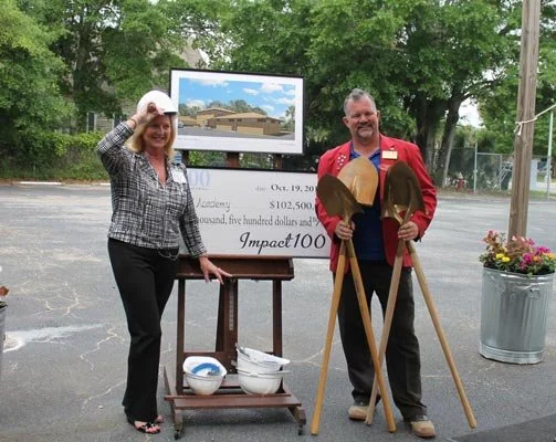 Two people standing outdoors next to a sign and a large check, holding shovels, with trees and a parking lot in the background.