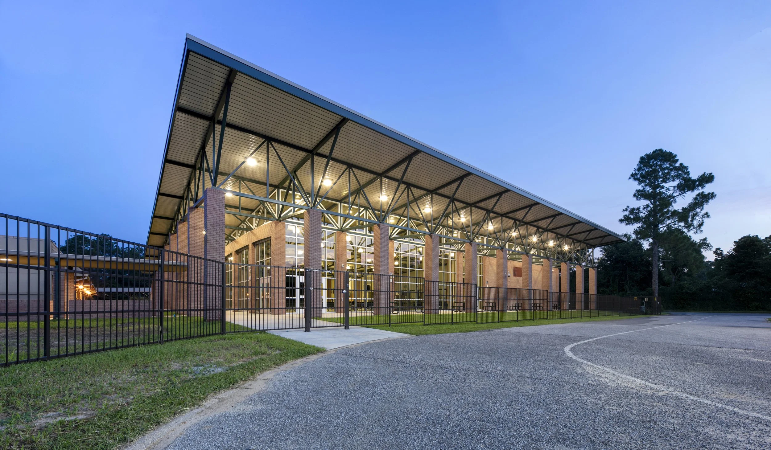 A school cafeteria with a metal and brick exterior, large glass windows, and a cantilevered roof, surrounded by a black metal fence and trees, illuminated at dusk.