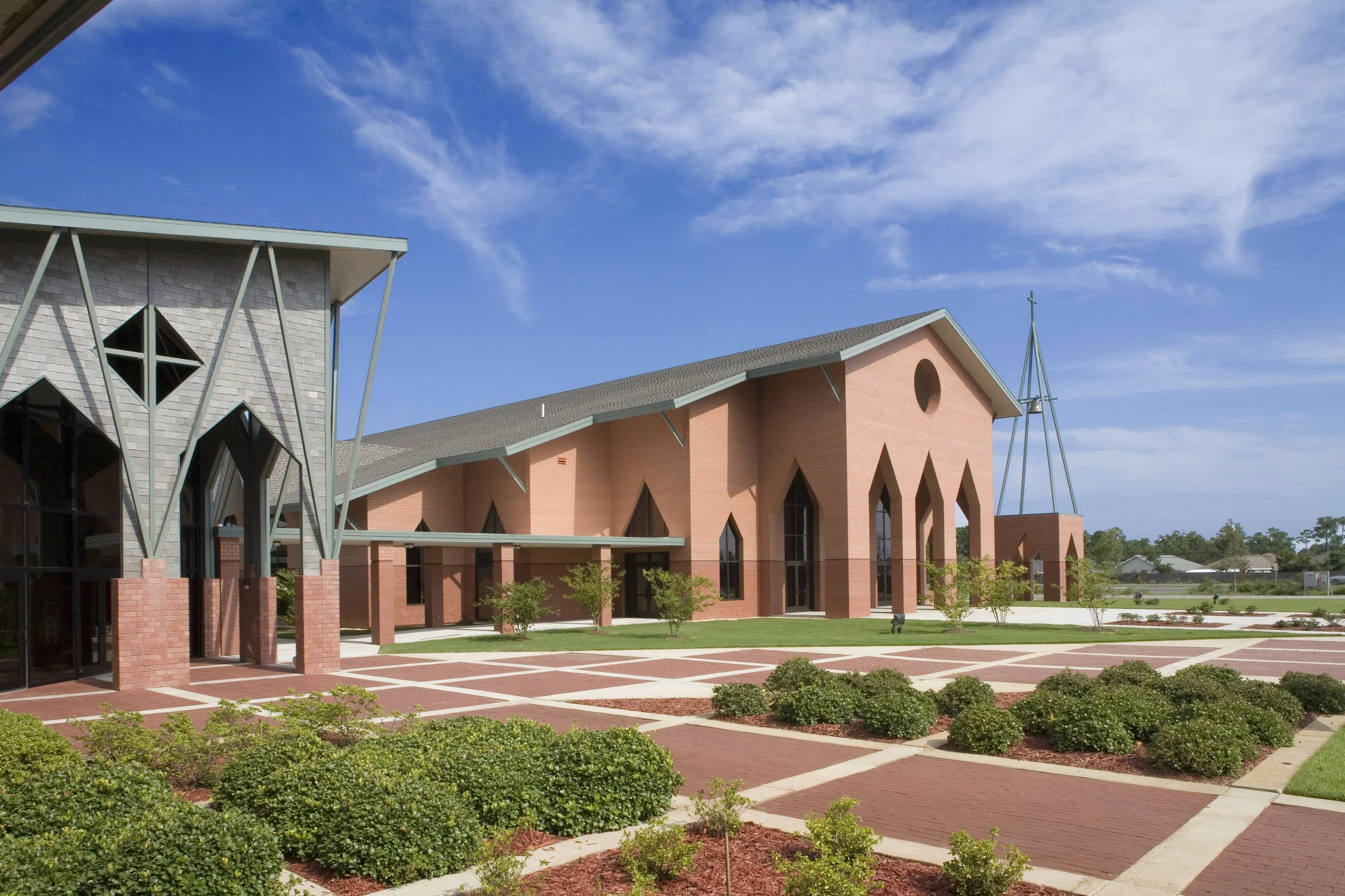 Colorful tall pink red brick church building under a bright blue sky with scattered clouds, surrounded by well-maintained landscaping and paved walkways.