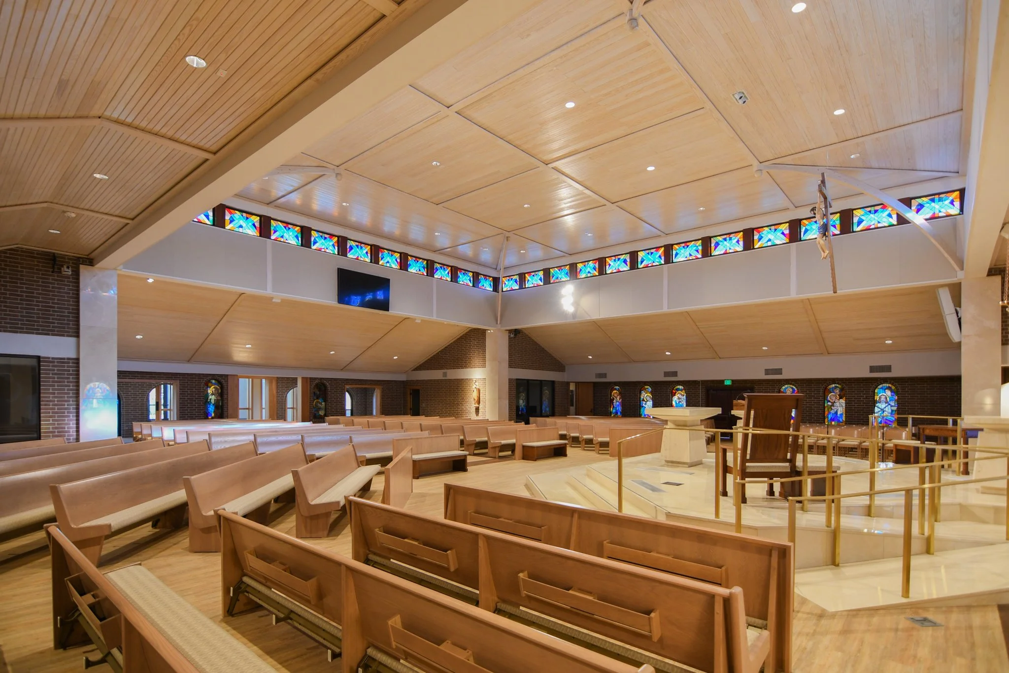 Interior of a church or chapel with wooden pews, stained glass windows, and a wooden ceiling with small spotlights.