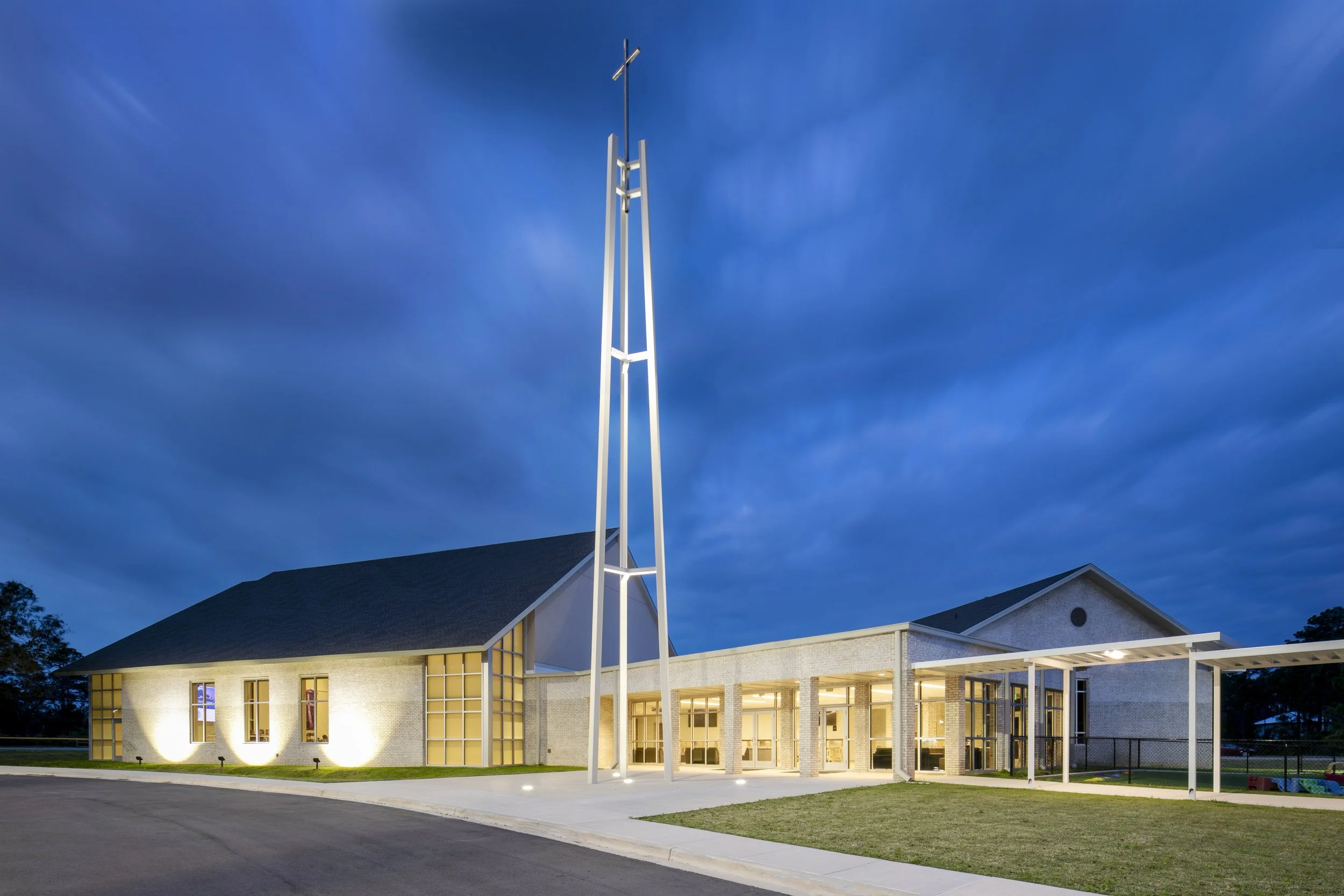 A modern church building at dusk with illuminated exterior lights. The church features large glass windows, a pitched roof, and a tall steeple with a cross on top. The sky is a dark blue with some clouds.