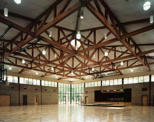 Large indoor gymnasium with brown ceiling beams and multiple hanging lights, featuring a stage at the far end and large glass windows along the top of the walls.
