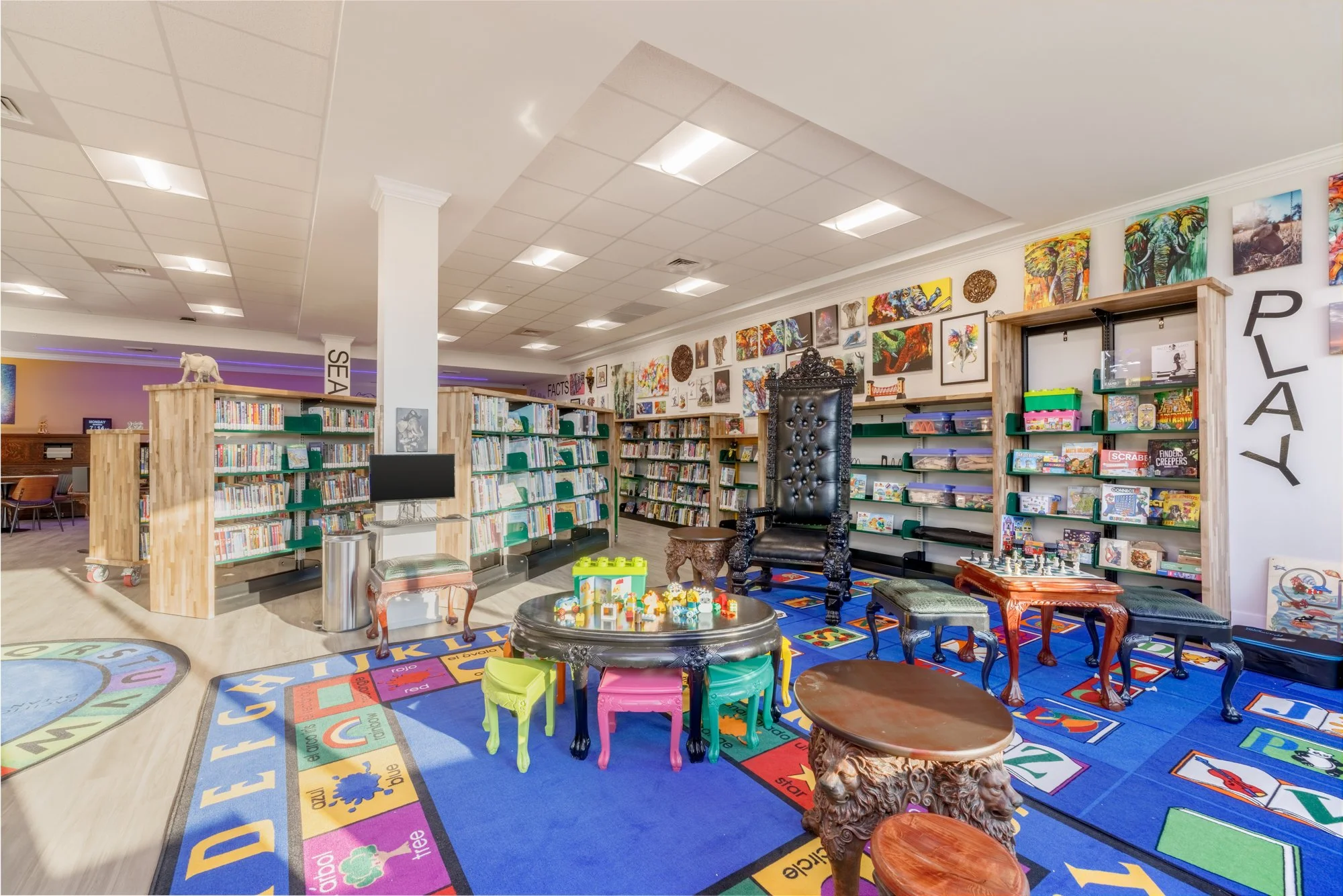 A children's play and reading area inside a library with bookshelves, colorful chairs, and educational toys on a patterned rug with alphabet and word labels.
