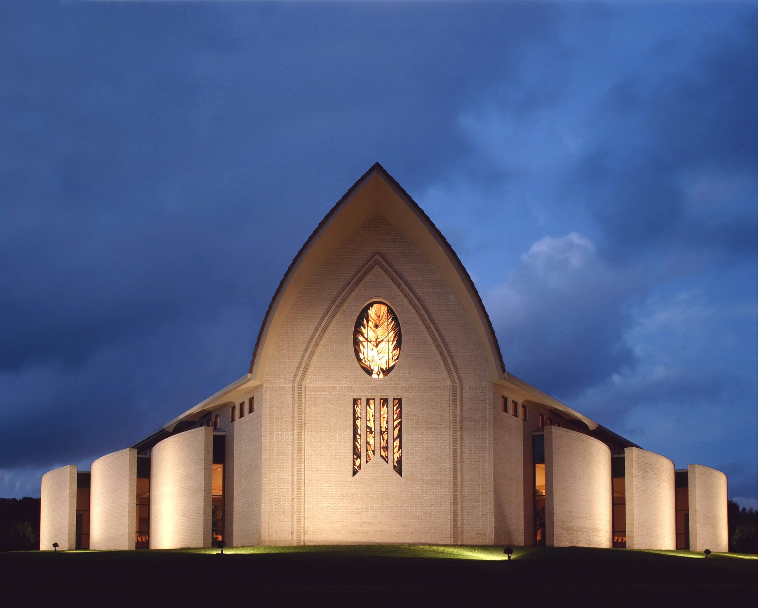 A church building illuminated at night with a pointed roof, arched stained glass window, and curved architectural walls, under a blue cloudy sky.
