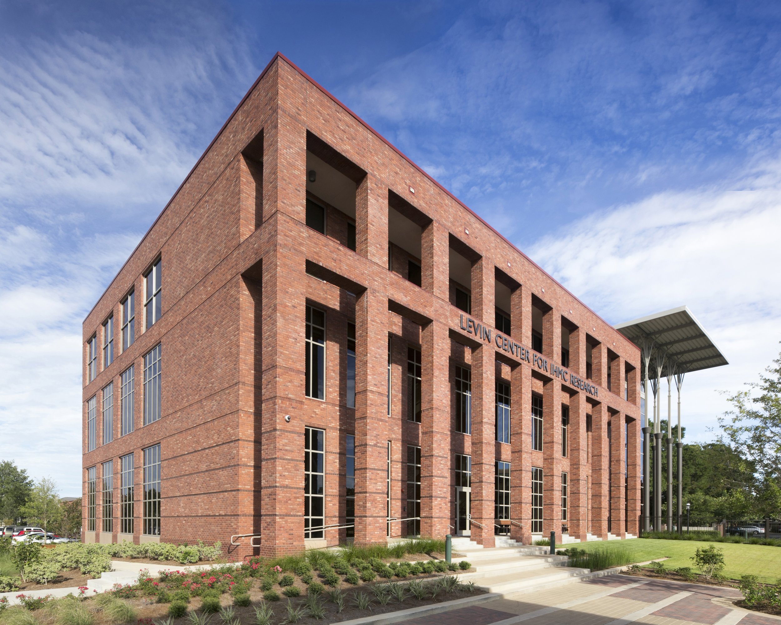 A three story modern brick building with multiple windows and a sign that reads 'Levin Center for IHMC Research.' The building has a distinctive corner with an overhang and is surrounded by landscaped gardens and a parking lot.