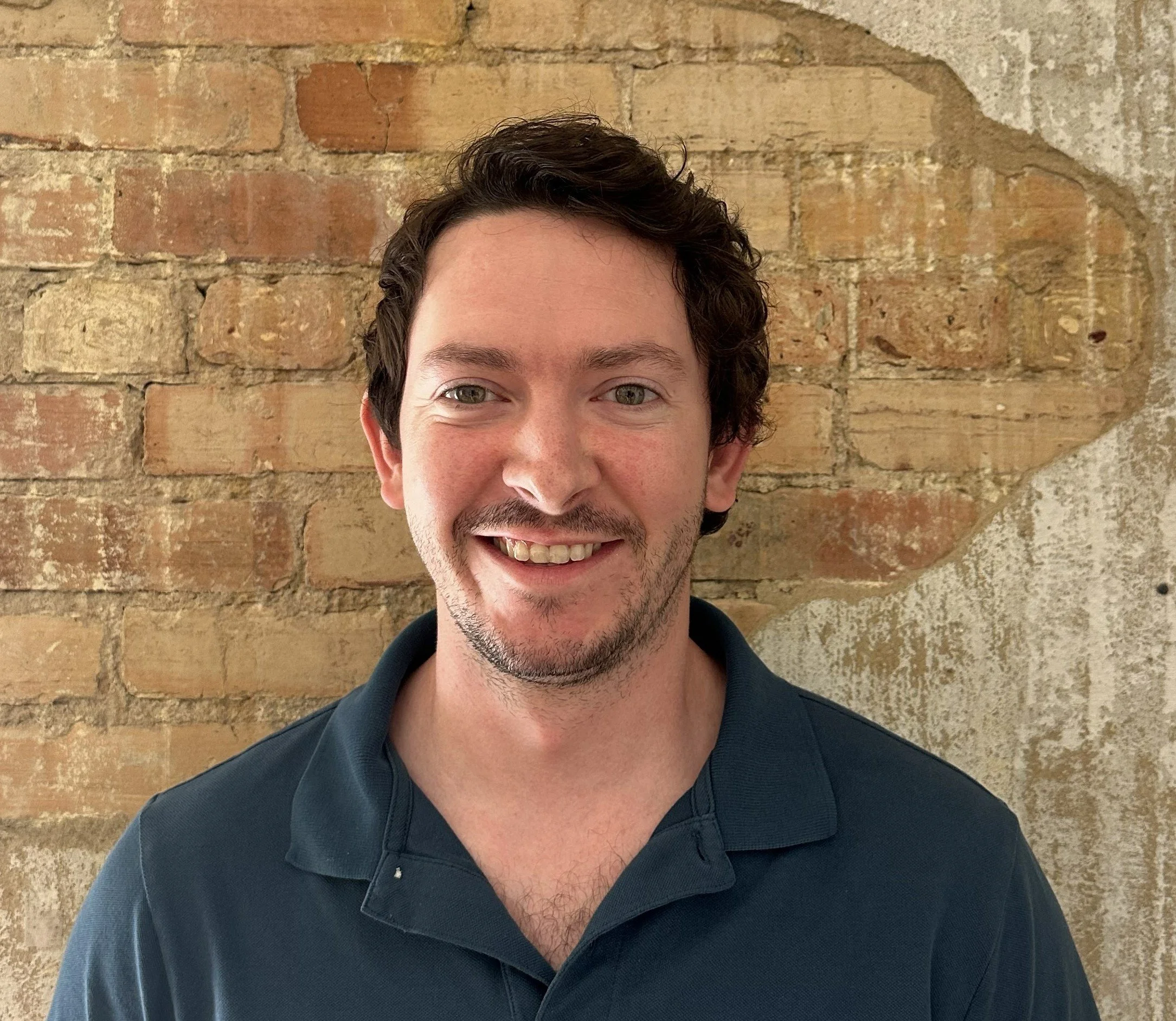 A smiling young man with short dark curly hair and light skin, wearing a dark blue polo shirt, standing in front of a brick wall.