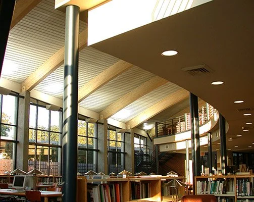 Interior view of a school library with large windows, high arched ceiling and beams, with bookshelves.