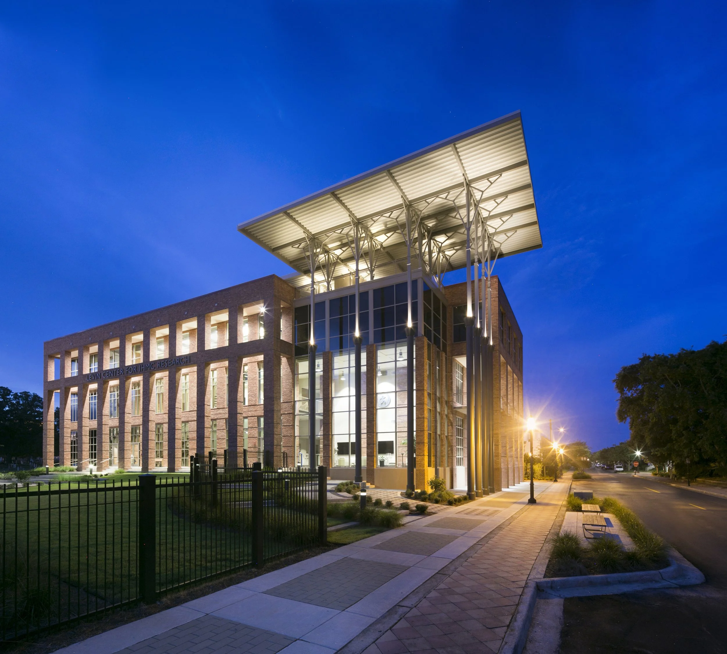 Night view of the Levin Center for IHMC Research building with illuminated interior and exterior lighting, a modern architectural design with glass and brick facade, and an overhanging roof structure with supporting columns, set against a dark evenin