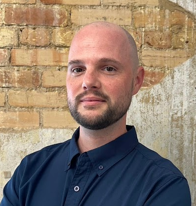 A man with a bald head and beard wearing a navy blue shirt, standing in front of a brick wall.