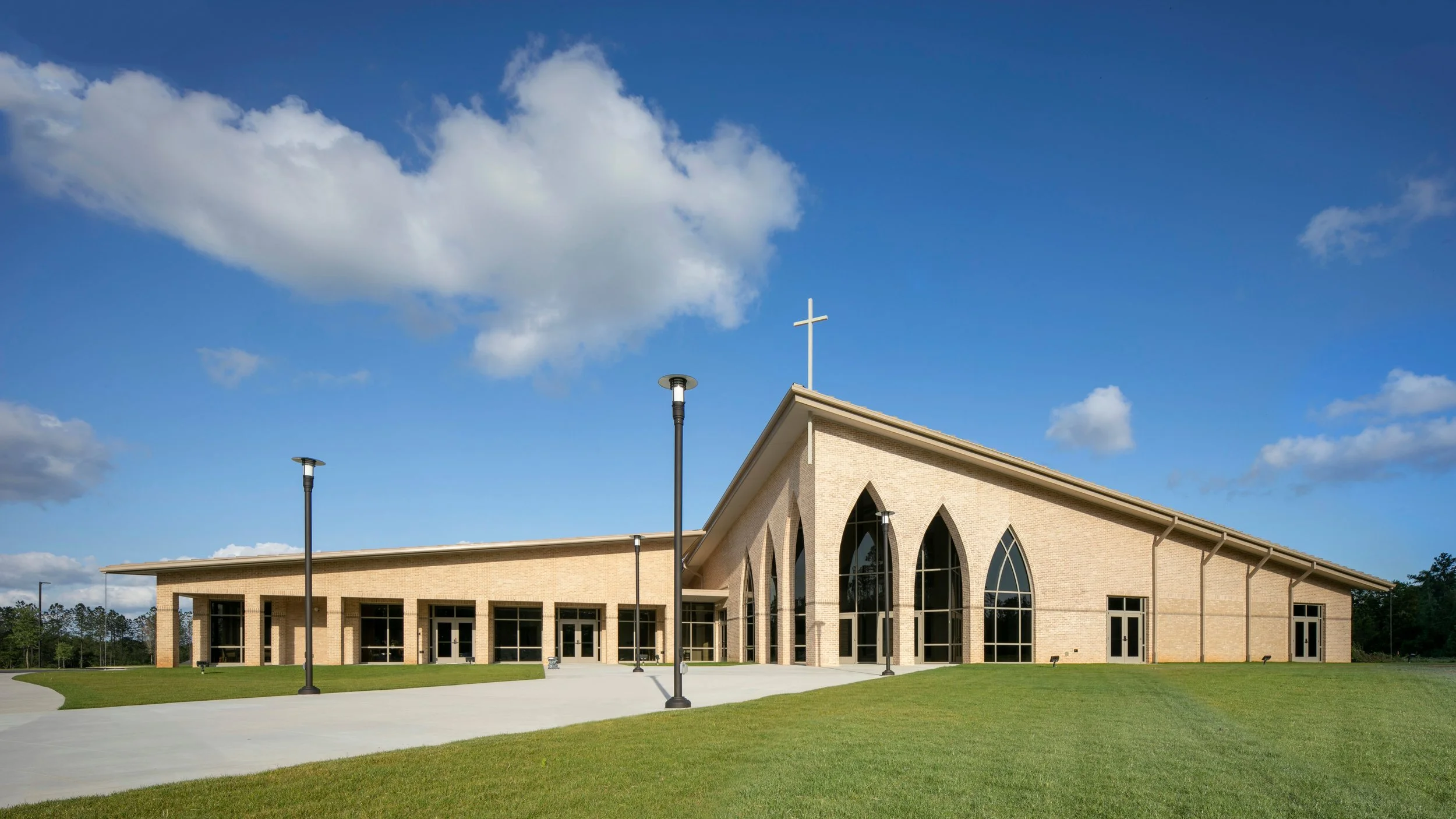 A modern tan brick church building with large pointed arched windows and a cross on top, set against a blue sky with scattered clouds.