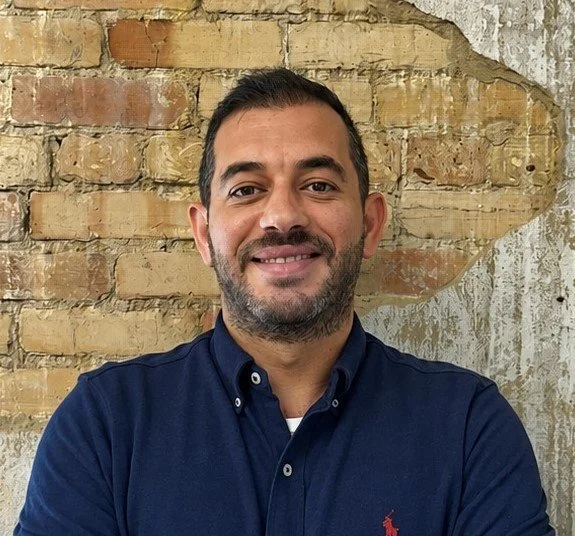 A man with dark short hair and a beard smiling at the camera, wearing a navy blue shirt, standing against a brick wall.