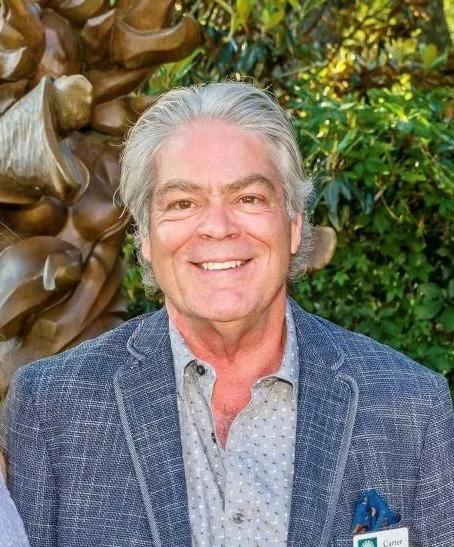 A smiling man with gray hair, wearing a gray blue blazer and a light patterned shirt, standing outdoors with green foliage and a palm tree in the background.