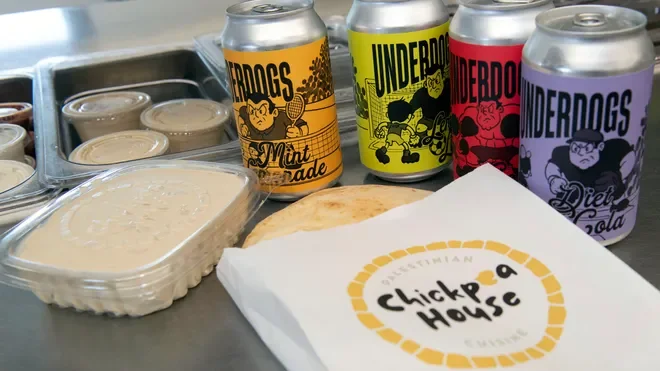 Four cans of soda in orange, yellow, red, and purple with a cartoon dog logo, along with a plastic container of hummus, a wrapped pita bread, and a paper bag with 'Chickpea House' logo, on a kitchen counter.