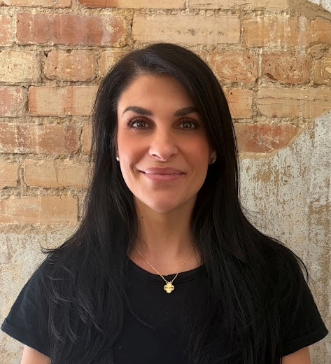 A woman with long black hair smiling, wearing a black shirt and a gold necklace, standing in front of a brick wall.
