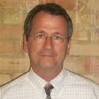 Portrait of a man with round glasses, wearing a light-colored button shirt and a patterned tie, standing in front of a brick wall.