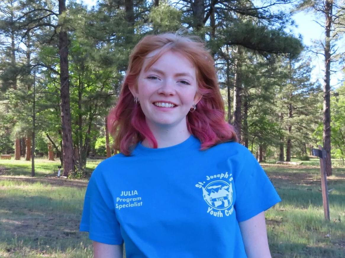 A young woman with blonde hair and a light-colored T-shirt standing outdoors in a wooded area with tall trees and sunlight.