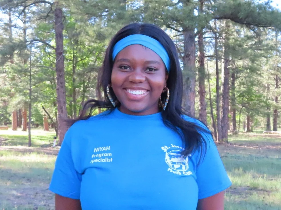 Young woman standing outdoors in a wooded area, smiling at the camera. She has dark hair, wears a white headband and a light blue t-shirt with logos and text, including 'NIYAH Program Specialist' and 'St. Joseph's Youth Camp'.
