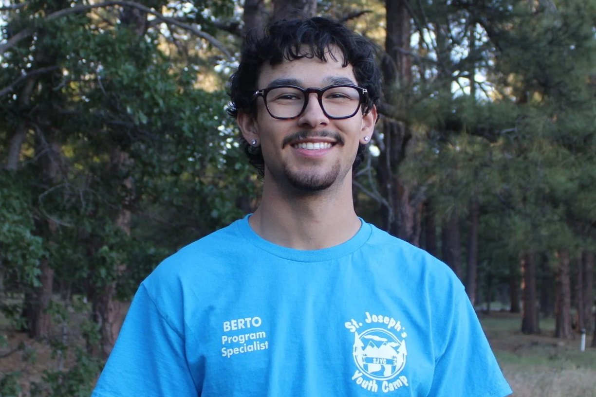 Young man with dark curly hair and a mustache, smiling, standing outdoors in a wooded area with green trees and grass.
