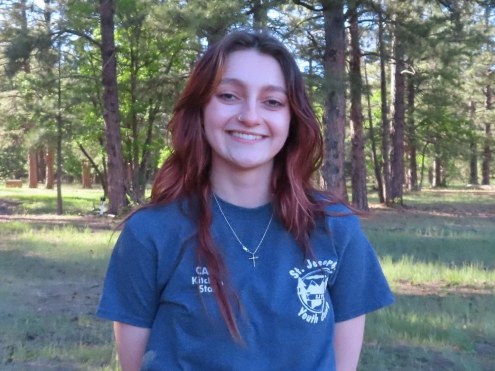 Young woman with red hair in braids, smiling and standing outdoors in a park with trees and grass.