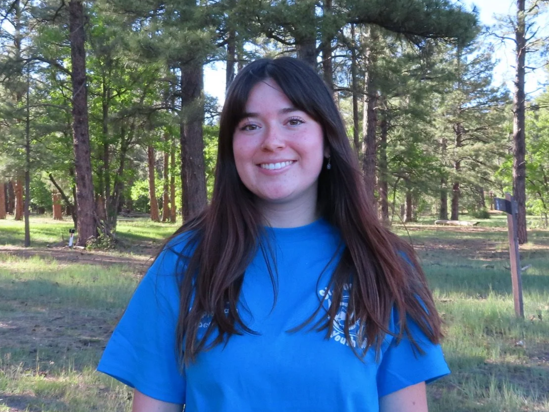 Young woman smiling outdoors in a wooded area, wearing a light grey t-shirt with printed text
