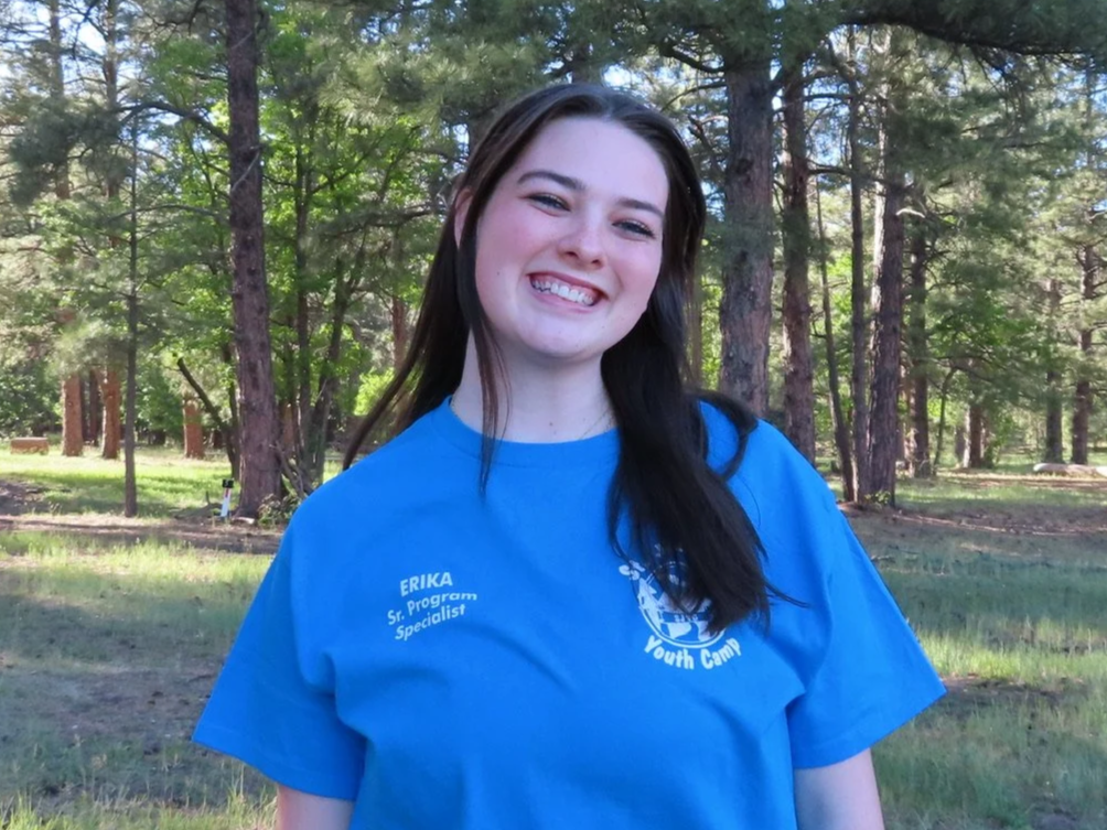 A smiling young woman with long brown hair standing outdoors in a wooded area with tall trees in the background, wearing a light blue t-shirt that has the words "ERIKA Program Specialist" and a logo on it.