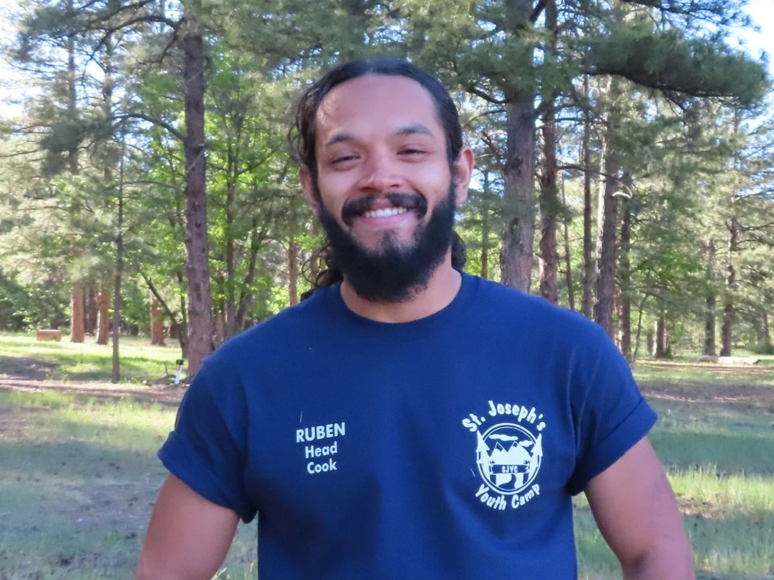A man with long curly hair and a beard smiling with his arms crossed, wearing a navy blue t-shirt in a wooded outdoor setting.