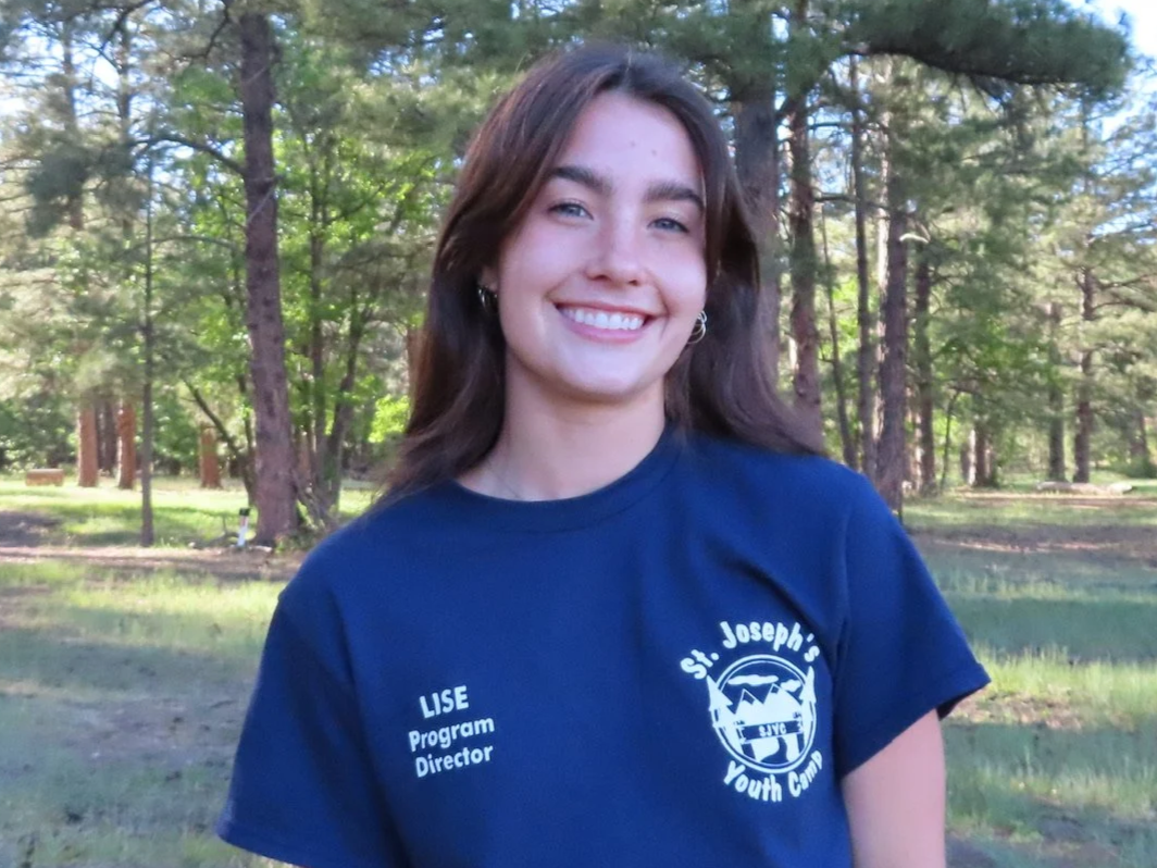 Young woman smiling outdoors in a forested area, wearing a light blue camp T-shirt with her name and role printed on it, and gold earrings.