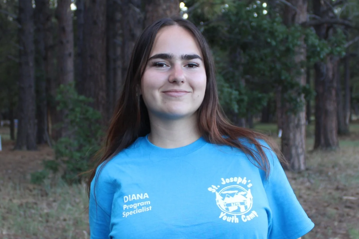 Young woman smiling outdoors in a wooded area, wearing a dark gray t-shirt with text that includes "Diana" and "Kitchen Staff" on the left chest and a camp logo on the right chest.