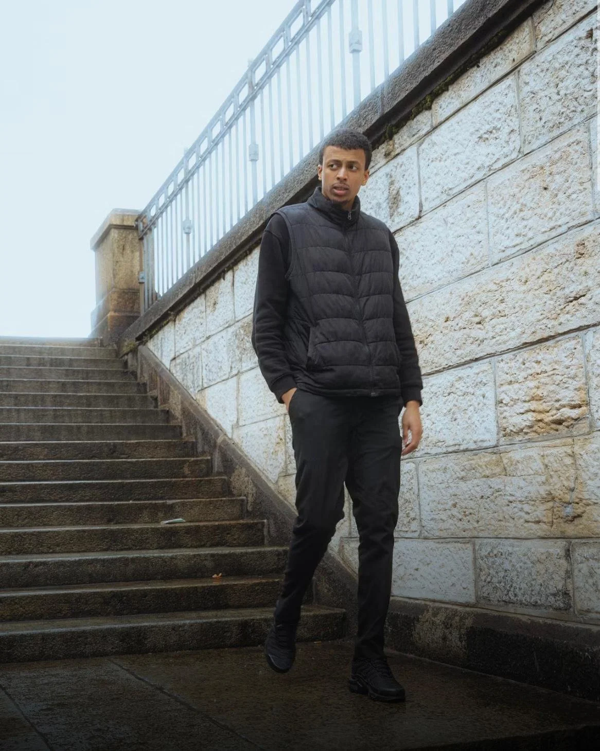 A young man dressed in black, walking down steps along a stone wall with a metal railing, on a cloudy day.