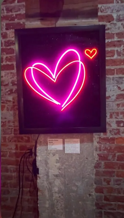 Neon artwork of two intertwined hearts, one larger pink and one smaller orange, on a black background, mounted on a brick wall.