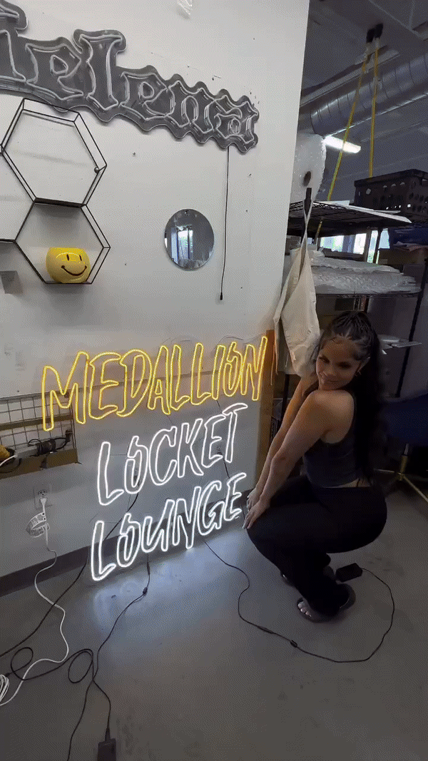 A woman sitting on the floor in front of a neon sign that reads 'MEDALLION LOCKET LOUNGE' with decorative items on the wall and an industrial-style interior in the background.