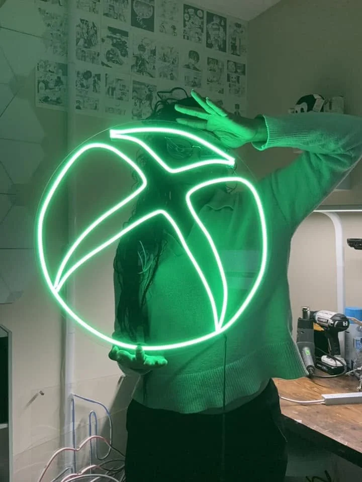 Person taking a mirror selfie with a neon green Xbox logo sign, obscuring part of their face. The background features a wall decorated with comic book art.